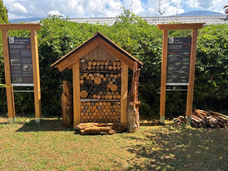 Holzhaus mit Giebeldach, dicht gefüllt mit gestapelten Holzscheiben, zwischen zwei Info-Tafeln des Lehrpfads