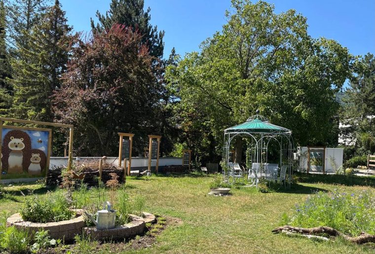 Kräuterspirale aus Steinen im Vordergrund, dahinter Pavillon und bunte Info-Tafel des Lehrpfads