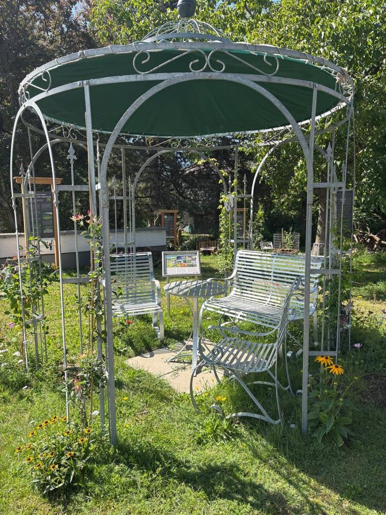 Pavillon des Lehrpfads von innen mit weißen Bänken, Tisch und Info-Tafel, umgeben von blühenden Stauden