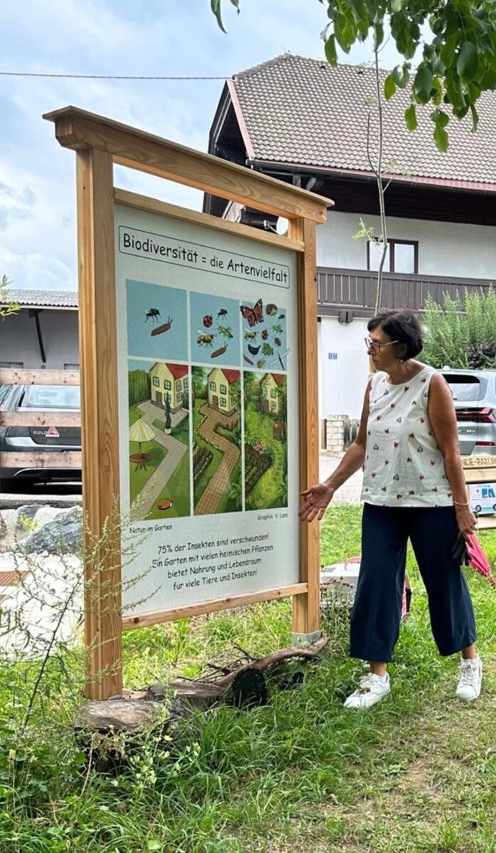 Info-Tafel „Biodiversität = die Artenvielfalt“ im Holzrahmen auf dem Lehrpfad, davor steht Ursula Ertl und zeigt auf die Tafel