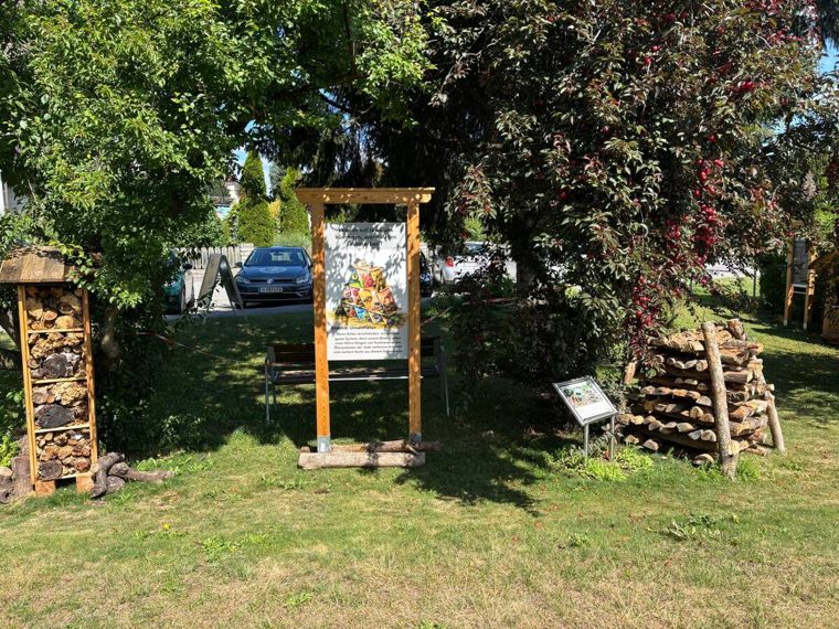 Lehrtafel in Holzrahmen auf der Wiese, links ein Insektenhotel, rechts ein Holzstapel und eine kleine Pulttafel