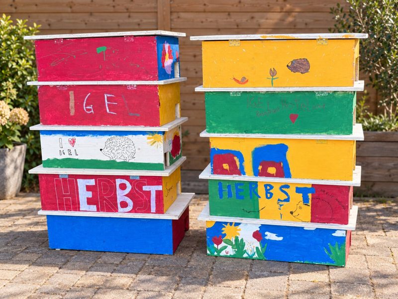 Zwei Stapel bunt bemalter Igel-Futterhäuser aus Holz, von Kindern mit Igeln, Blumen und Schriftzügen gestaltet