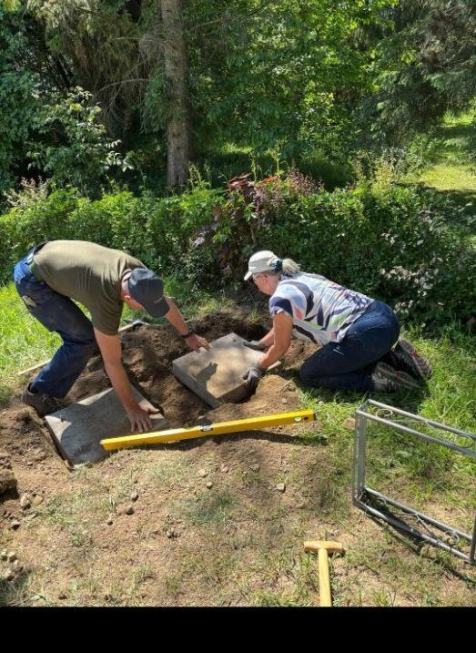 Zwei Helfende legen knieend eine Betonplatte als Fundament in die ausgehobene Erde und richten sie mit einer gelben Wasserwaage aus