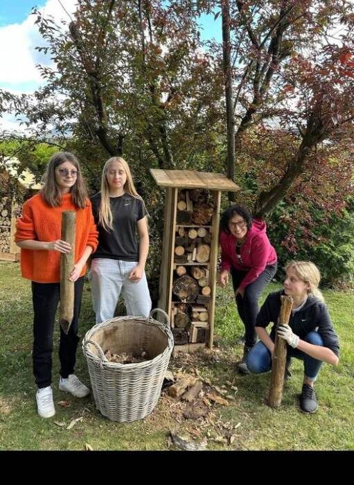 Vier Jugendliche stehen mit Holzstämmen und einem Weidenkorb neben dem fertigen Insektenhotel aus Holz, gefüllt mit Rundhölzern und Zapfen