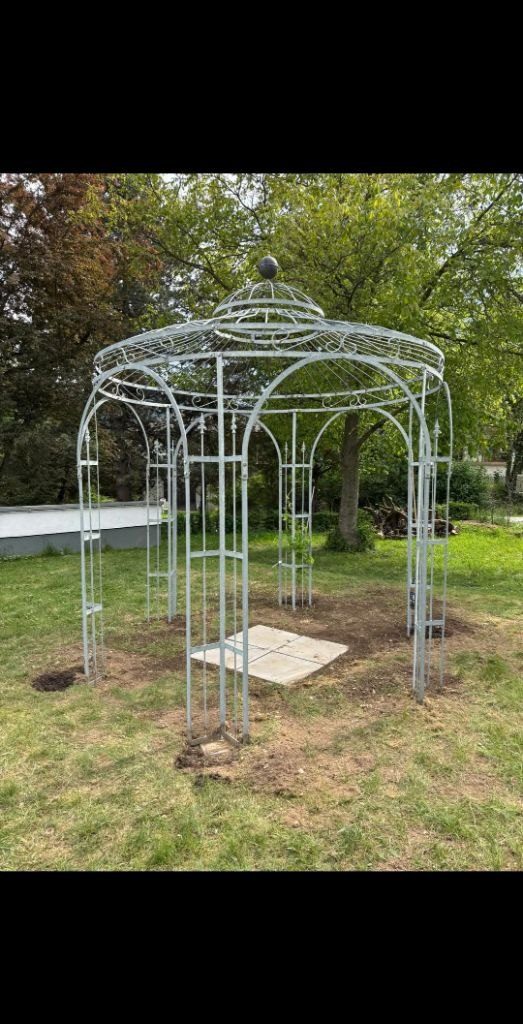 Fertig aufgestellter Pavillon aus hellgrauem Ziergitter-Metall mit gewölbtem Dach auf der Wiese, darunter eine Steinplatte als Boden
