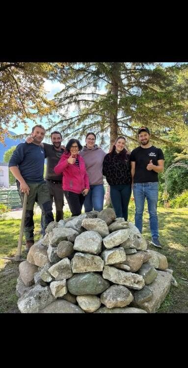 Sechs Helferinnen und Helfer stehen mit erhobenen Daumen hinter der fertig aufgeschichteten Steinpyramide aus großen Flusssteinen unter einer Fichte